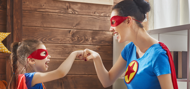 Woman and girl in red superhero masks and capes fist-bump in a cozy room with a wooden wall and star pillow nearby.