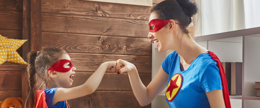 Woman and girl in red superhero masks and capes fist-bump in a cozy room with a wooden wall and star pillow nearby.