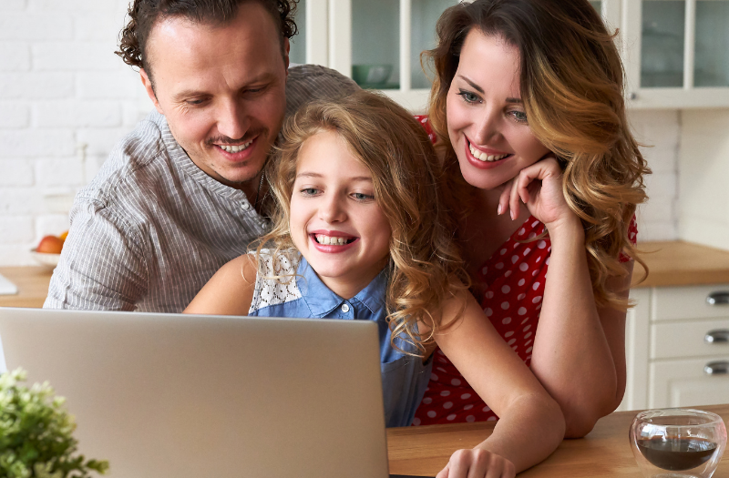 Family of three gathered around a laptop at a kitchen island, smiling at the screen