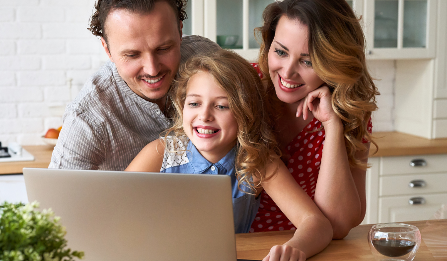 Family of three gathered around a laptop at a kitchen island, smiling at the screen