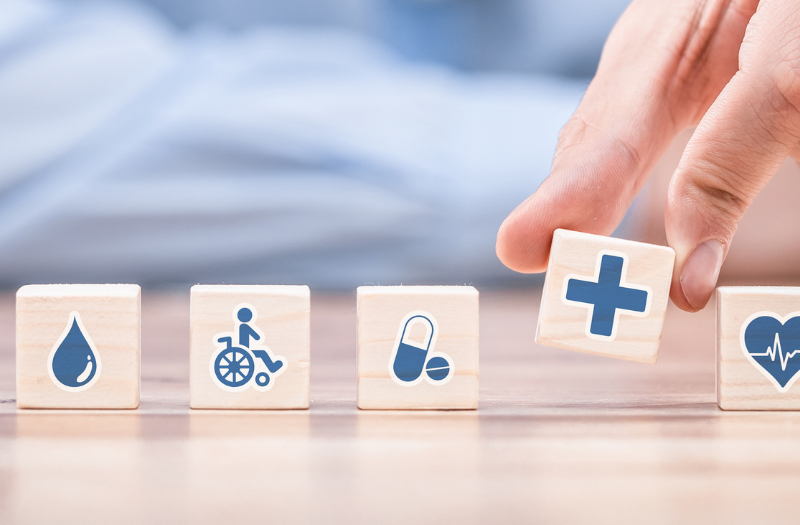 Row of wooden blocks with health icons: blood drop, wheelchair, capsule, then a hand placing a block with a plus sign, followed by a heart with a heartbeat line on the last block.