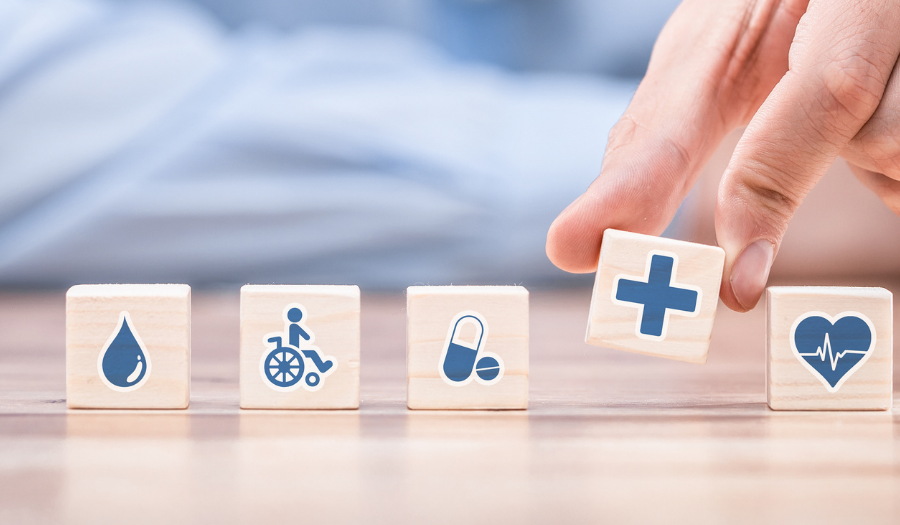 Row of wooden blocks with health icons: blood drop, wheelchair, capsule, then a hand placing a block with a plus sign, followed by a heart with a heartbeat line on the last block.