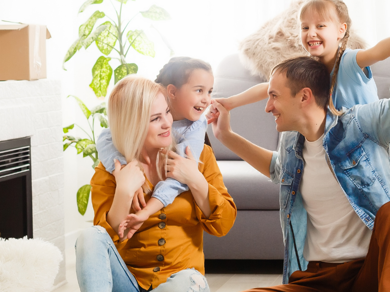 Family in a new home moment: parents with two kids celebrating amid moving boxes and packed belongings in a bright living room.