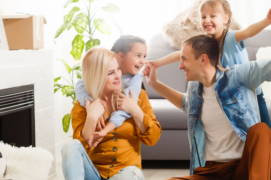 Family in a new home moment: parents with two kids celebrating amid moving boxes and packed belongings in a bright living room.