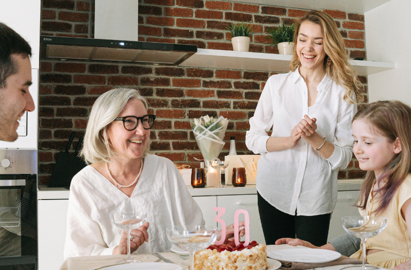A family gathers around a kitchen table for a birthday celebration, cake with pink '30' candles in front of them.