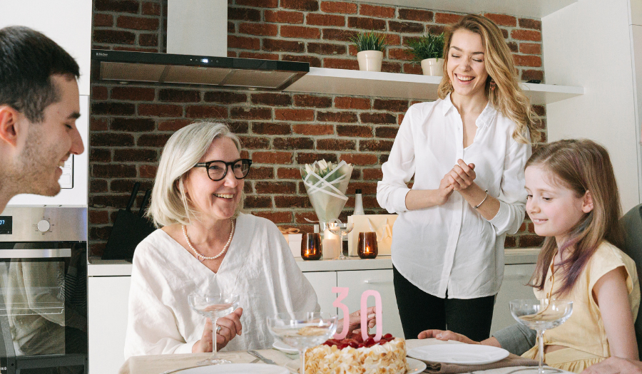 A family gathers around a kitchen table for a birthday celebration, cake with pink '30' candles in front of them.