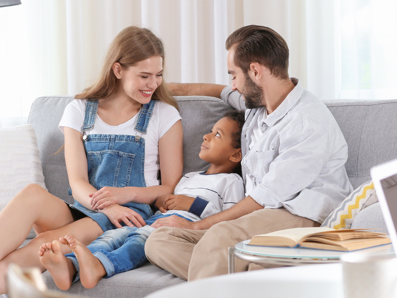 A happy family sits on a gray sofa—mother, father, and their child cuddled together in a bright living room.