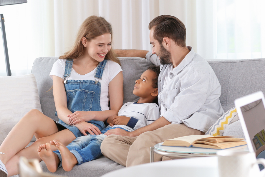 A happy family sits on a gray sofa—mother, father, and their child cuddled together in a bright living room.