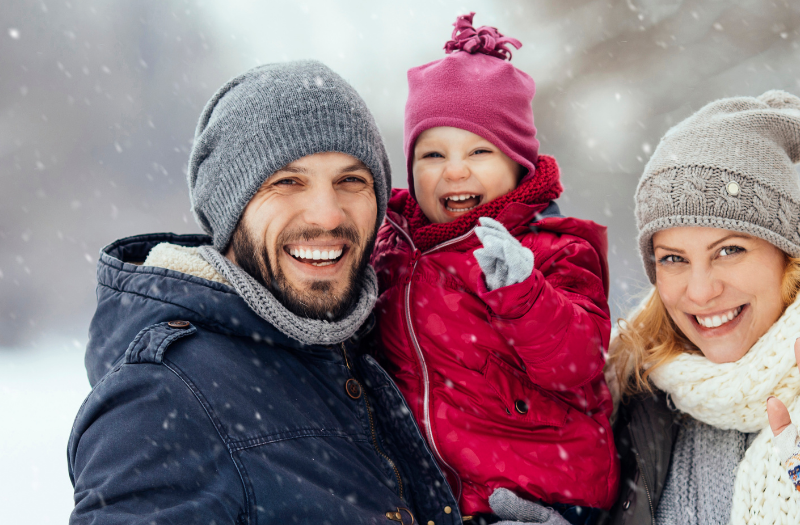 Cheerful family of three in winter coats posing in a snowy park; father holds a laughing child, while the mother waves at the camera.