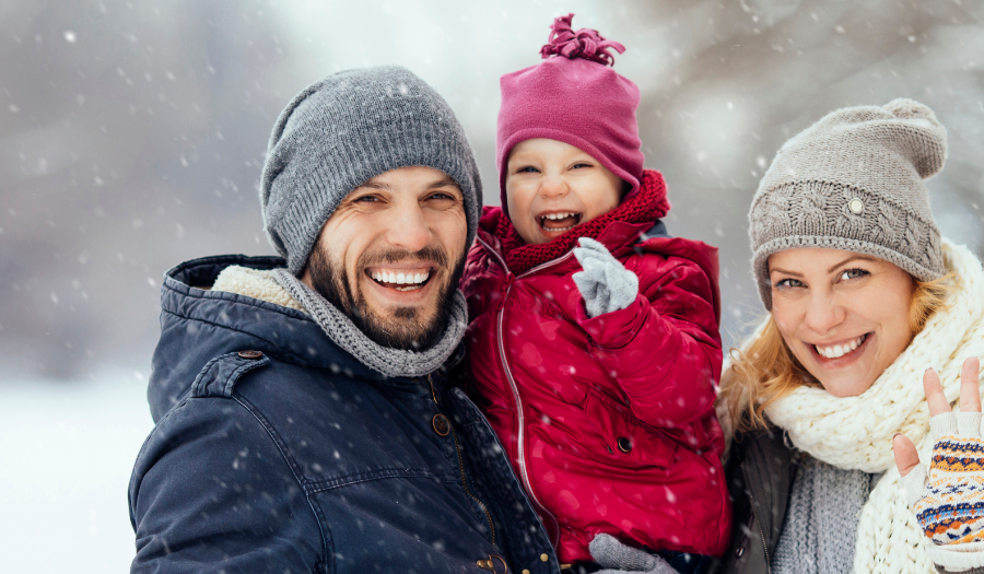 Cheerful family of three in winter coats posing in a snowy park; father holds a laughing child, while the mother waves at the camera.
