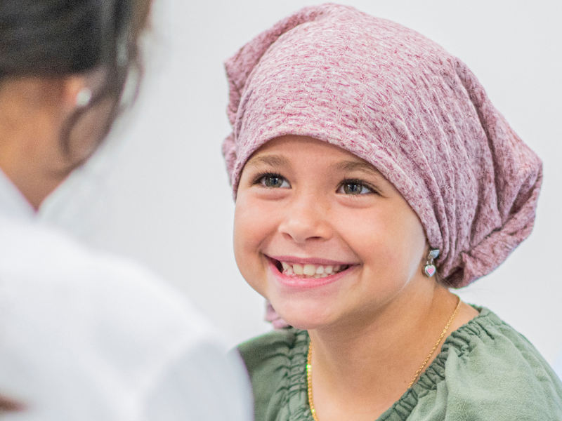 Young girl wearing a pink headscarf smiles at a healthcare professional during a medical visit outside a clinical setting? (informative alt)