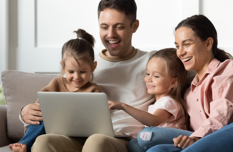 Family of four on a sofa, looking at a laptop together in a bright living room, smiling and interacting.