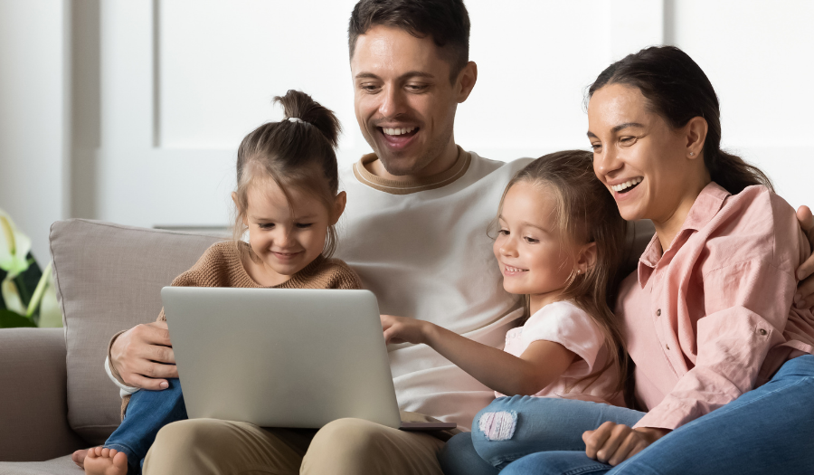 Family of four on a sofa, looking at a laptop together in a bright living room, smiling and interacting.