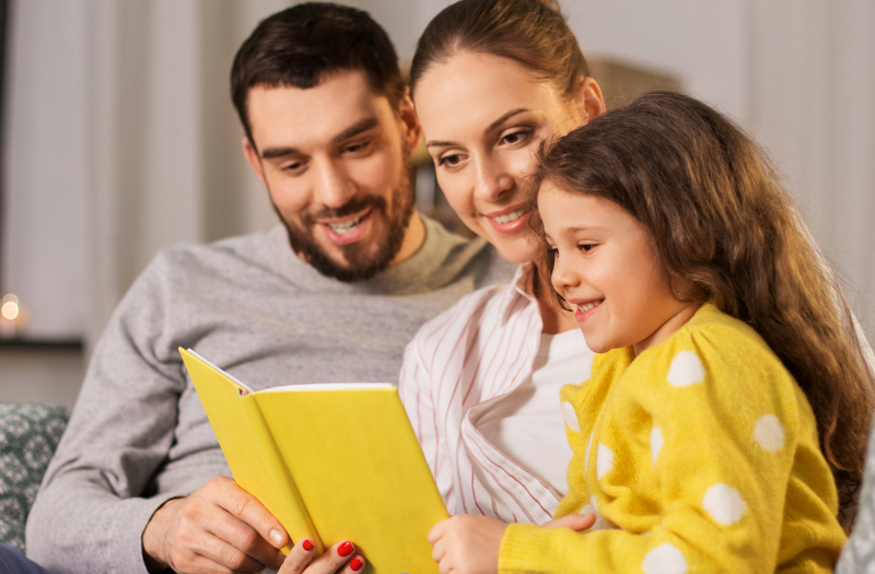 Family of three reading a yellow book together on a couch, smiling at the story.