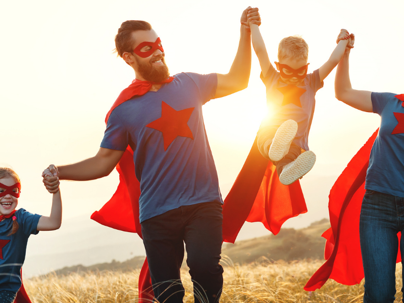 Family of four in matching blue shirts with red capes and masks, holding hands and lifting their youngest in a sunlit field at sunset.