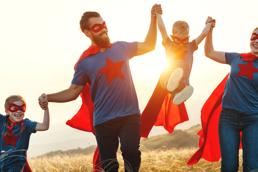 Family of four in matching blue shirts with red capes and masks, holding hands and lifting their youngest in a sunlit field at sunset.