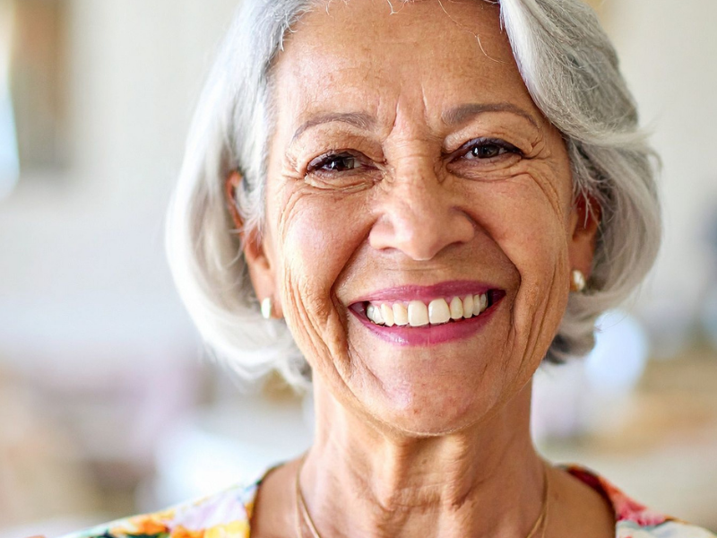 Smiling elderly woman with short gray hair in a bright indoor setting wearing a floral top.