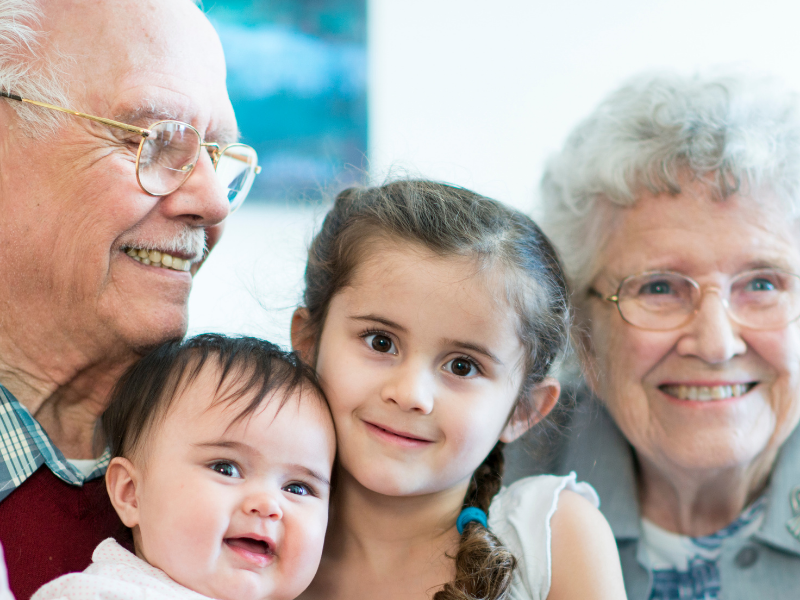 Grandfather, grandmother, and two young grandchildren smiling together on a sofa.7,?