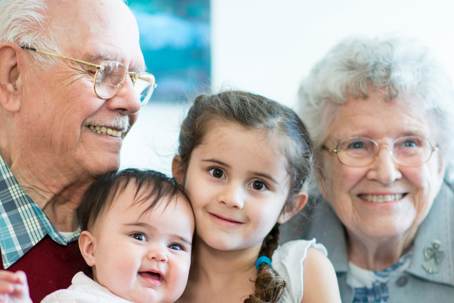 Grandfather, grandmother, and two young grandchildren smiling together on a sofa.7,?