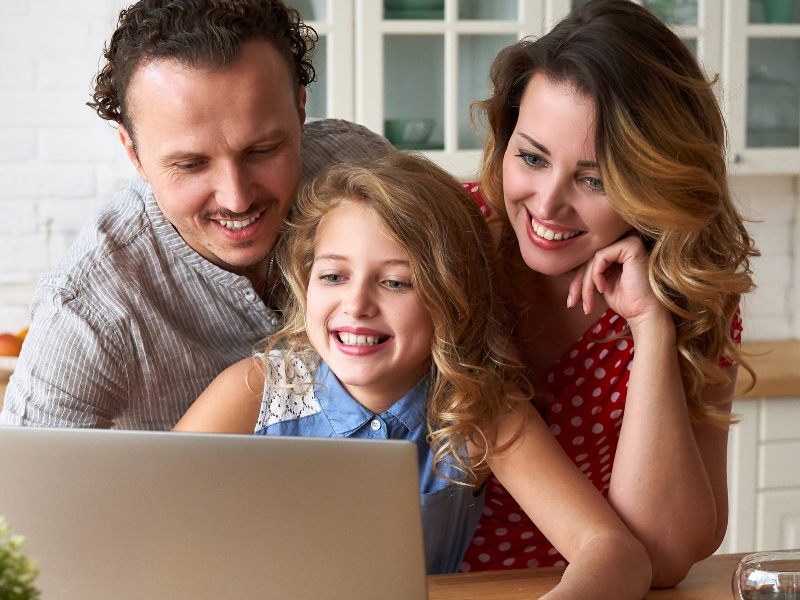 Family of three gathered around a laptop on a kitchen island, smiling at the screen with pink roses on the counter.