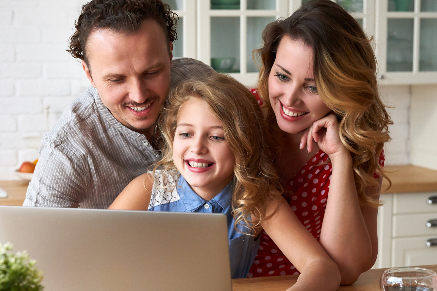 Family of three gathered around a laptop on a kitchen island, smiling at the screen with pink roses on the counter.