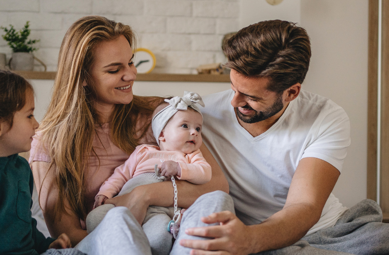 Mother and father sit on a bed with their baby girl in a headband, as the older sister watches and smiles in a cozy room with a child-friendly backdrop.