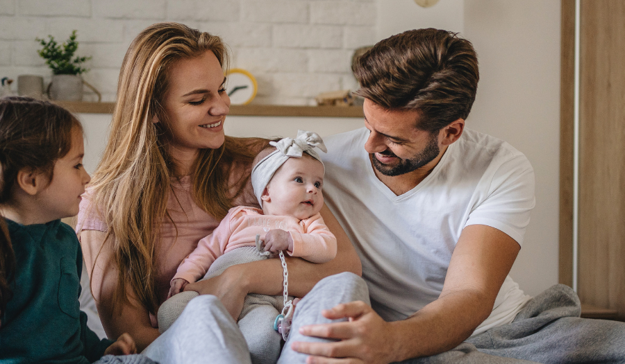 Mother and father sit on a bed with their baby girl in a headband, as the older sister watches and smiles in a cozy room with a child-friendly backdrop.
