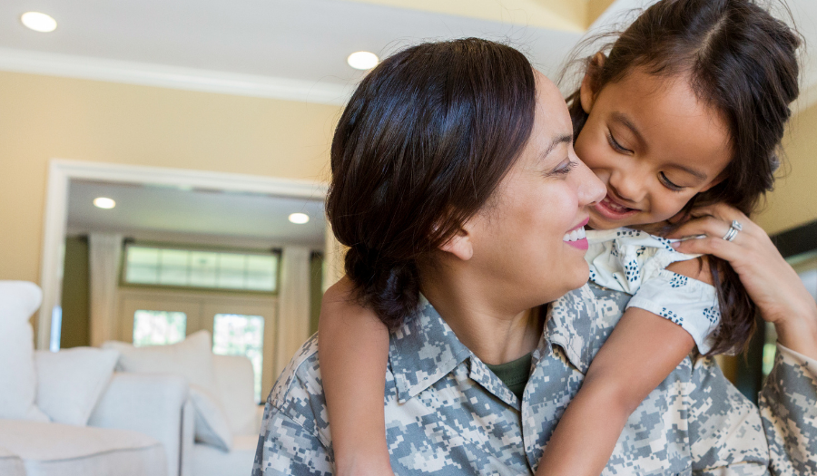 Soldier in camouflage uniform smiles as her young daughter hugs her from behind in a cozy living room.
