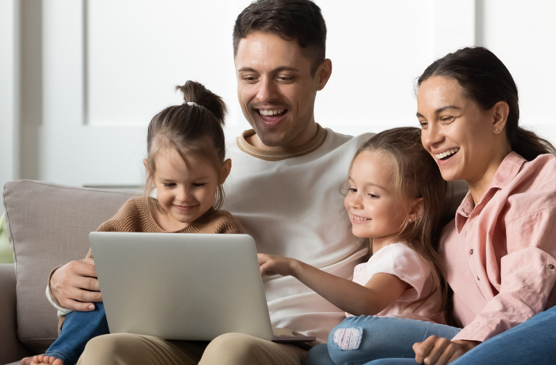 Family of four sits on a living room sofa, smiling at a laptop together on the dad's lap.