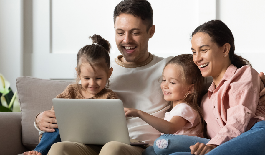 AccuQuote Blog 14 - AccuQuote Family of four sits on a living room sofa, smiling at a laptop together on the dad's lap.