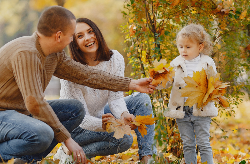 A couple kneels in a park covered in autumn leaves, smiling at their young daughter who collects yellow leaves with them.
