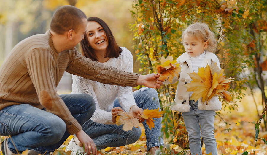 A couple kneels in a park covered in autumn leaves, smiling at their young daughter who collects yellow leaves with them.