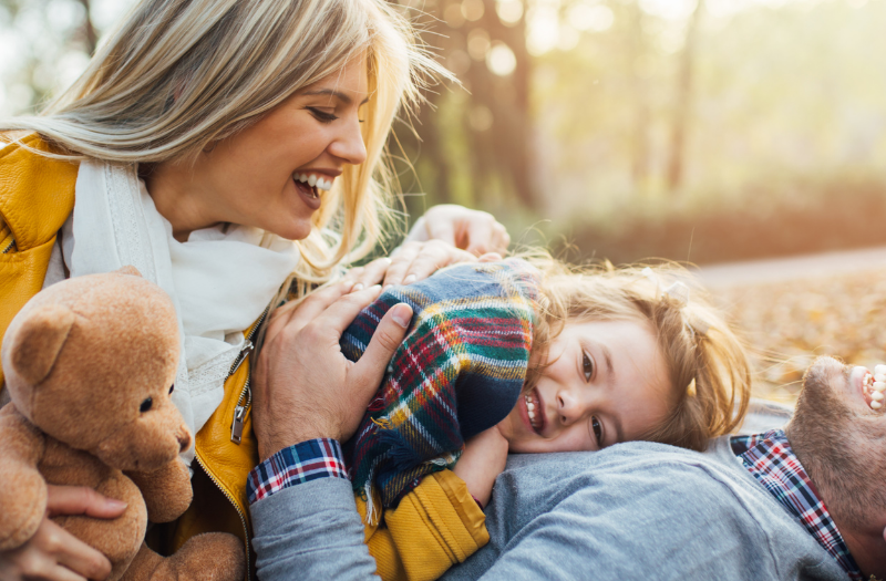Family enjoying an autumn day on a leaf-strewn ground; mom in a yellow jacket holds a teddy bear while dad and daughter cuddle and laugh together.