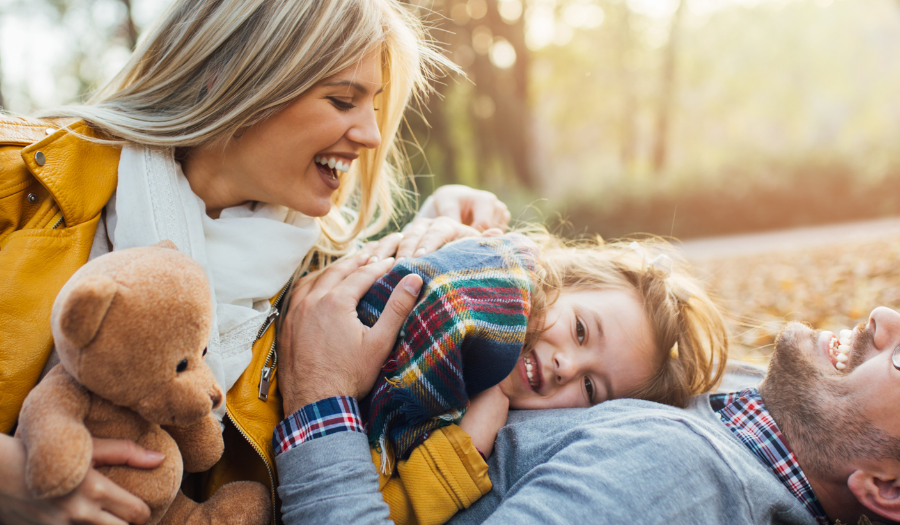 Family enjoying an autumn day on a leaf-strewn ground; mom in a yellow jacket holds a teddy bear while dad and daughter cuddle and laugh together.