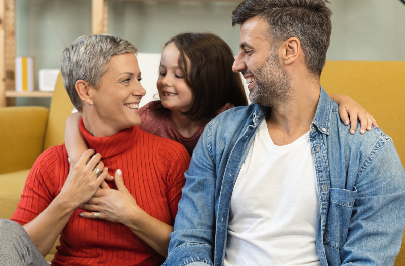 Joyful family moment on a yellow sofa: mother in a red sweater, father in a denim shirt, and child hugging them from behind.
