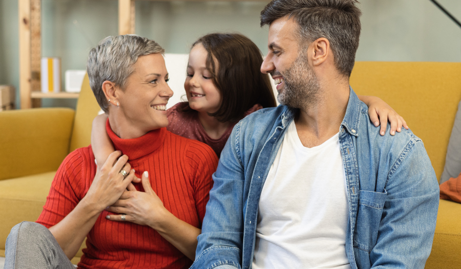 Joyful family moment on a yellow sofa: mother in a red sweater, father in a denim shirt, and child hugging them from behind.