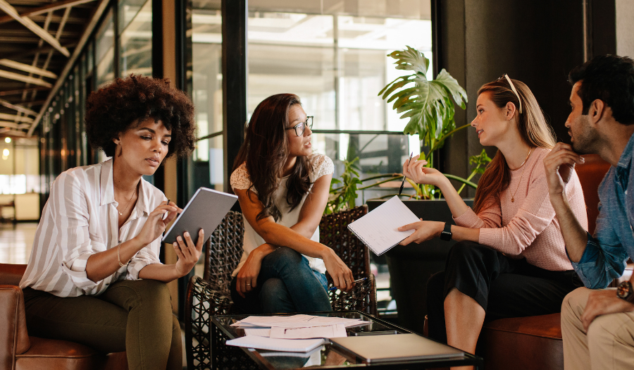 Four coworkers seated on couches in a modern office, discussing with a tablet and notebooks on a coffee table.