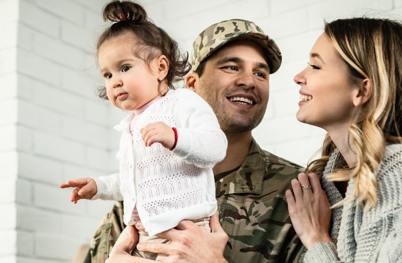 Soldier in camouflage holds a young child while partner smiles and touches his shoulder in a cozy living room setting.