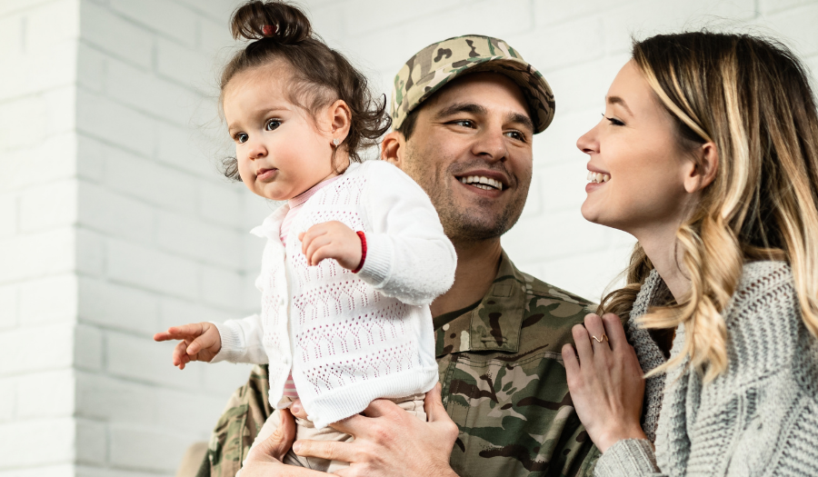 Soldier in camouflage holds a young child while partner smiles and touches his shoulder in a cozy living room setting.