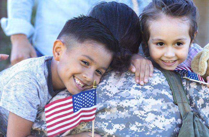 Two children hug a soldier in camouflage uniform, holding small American flags and a plush teddy bear.