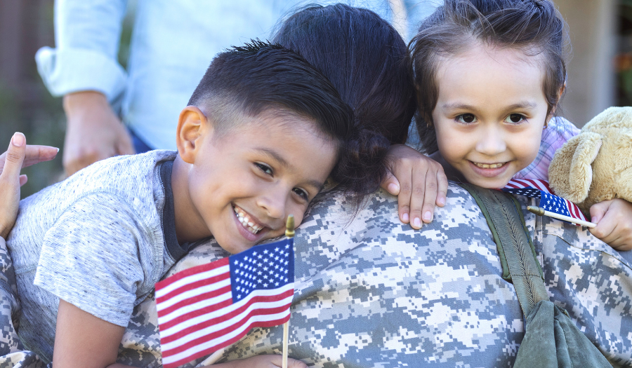 AccuQuote Blog 2 - AccuQuote Two children hug a soldier in camouflage uniform, holding small American flags and a plush teddy bear.