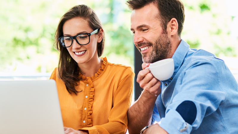 Two coworkers smiling at a laptop in a bright cafe, one holding a cup of coffee on the side of the desk.