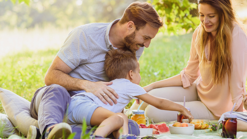 Family of four enjoying a sunny picnic on a blanket in a park, sharing fruit and drinks together.