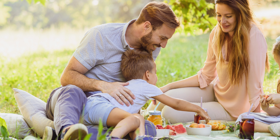 Family of four enjoying a sunny picnic on a blanket in a park, sharing fruit and drinks together.