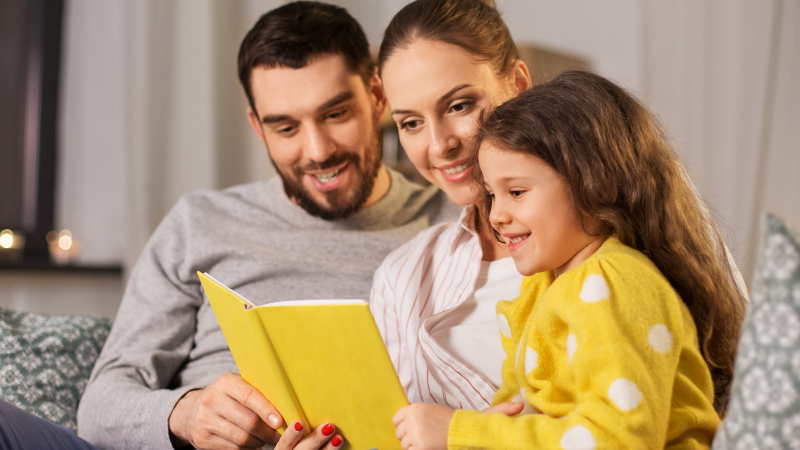 Family of three reading a bright yellow book together on a living room sofa.
