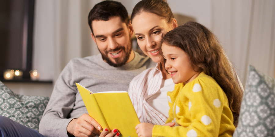 Family of three reading a bright yellow book together on a living room sofa.