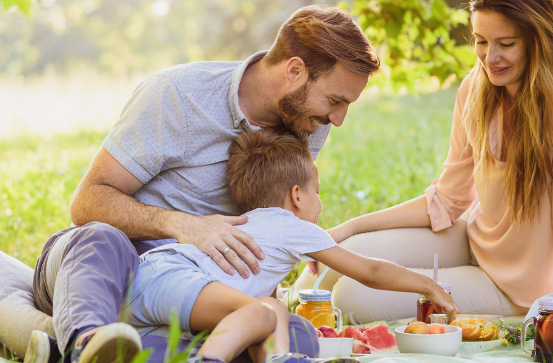 Family of four sharing a sunny outdoor picnic on a blanket in a park.