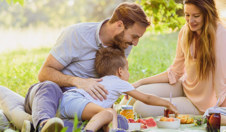 Family of four sharing a sunny outdoor picnic on a blanket in a park.