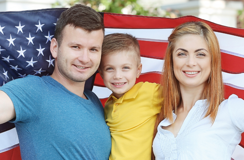 Family of three smiling, holding up a large American flag outdoors.