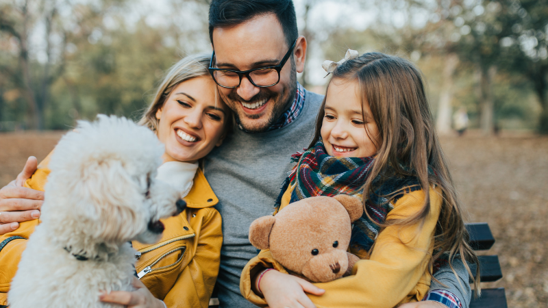 A smiling family sits on a park bench with a white fluffy dog and a teddy bear during autumn.